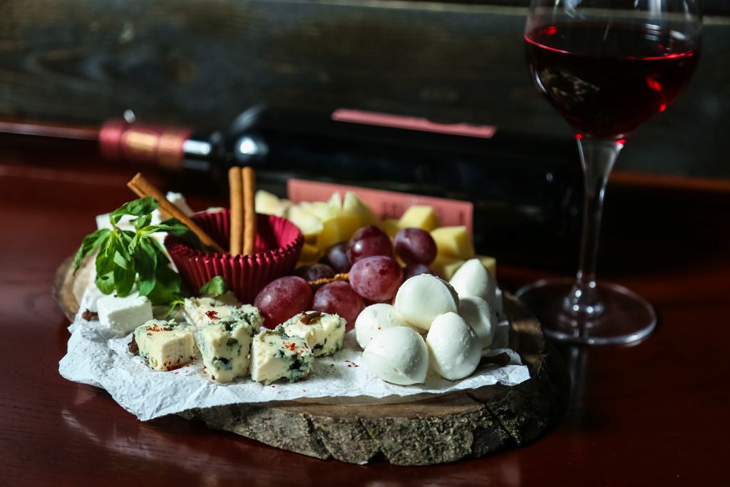 A plate of cheeses and grapes beside a glass and bottle of red wine