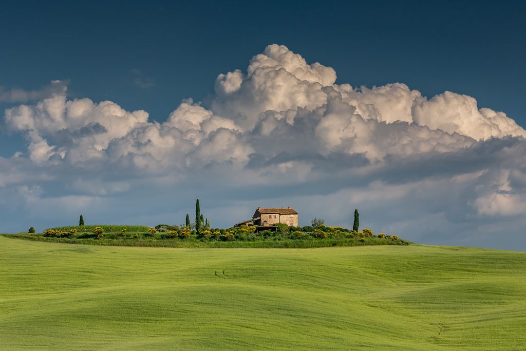 Stone farmhouse with cypresses on a green hill in Val d'Orcia under clouds