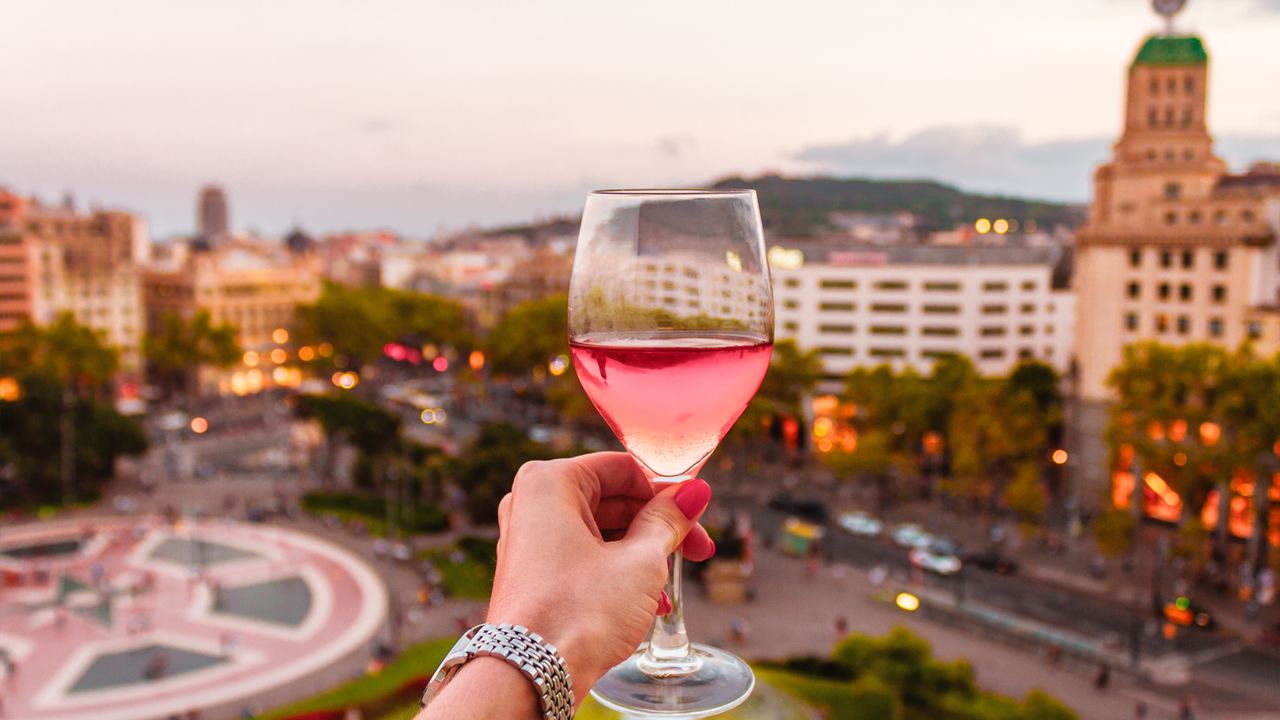 A hand holding a glass of rose wine above Plaça de Catalunya in Barcelona at sunset