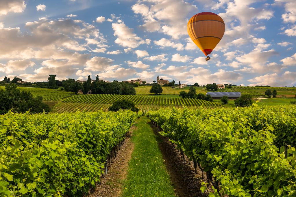 A colourful hot air balloon floating above rows of green vines with a village and church on the horizon in Bordeaux, France