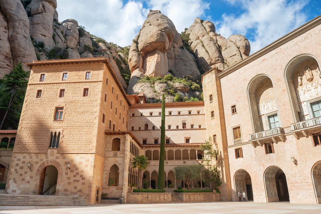 Courtyard of Santa Maria de Montserrat monastery with a cypress and rock formations behind