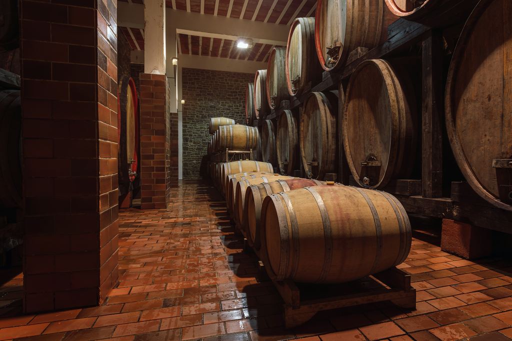 Rows of oak barrels lying on wooden cradles along a brick-floored wine cellar