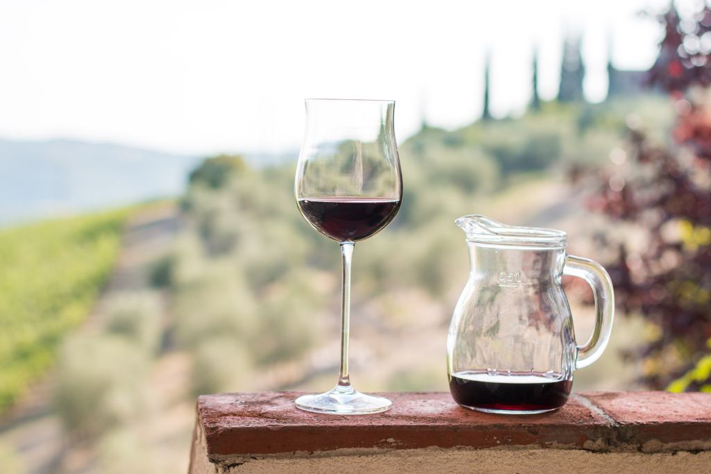 A glass and a carafe of red wine on a brick wall with Tuscan hills behind