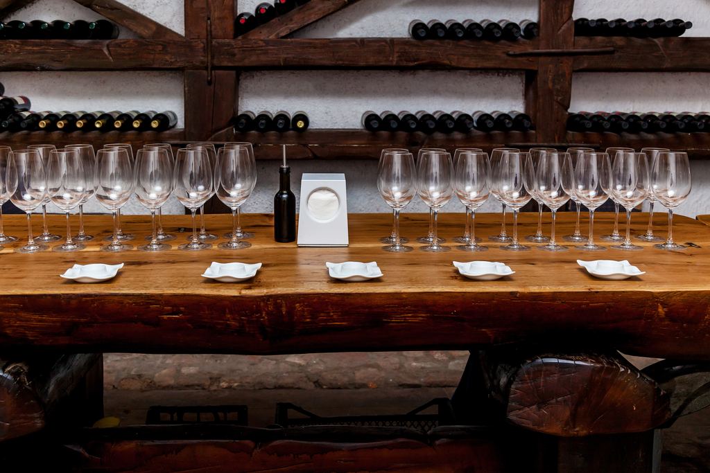 Wooden tasting table set with rows of empty wine glasses and small plates, with bottle racks behind