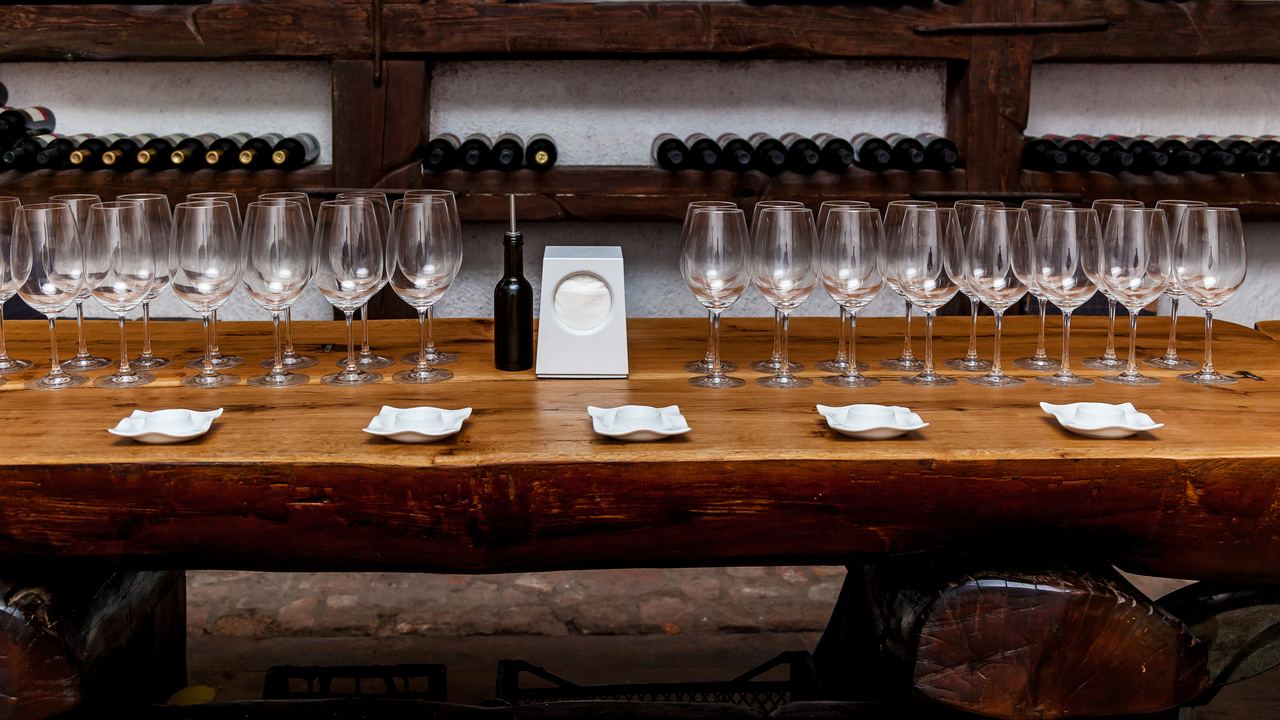 Wooden tasting table set with rows of empty wine glasses and small plates, with bottle racks behind
