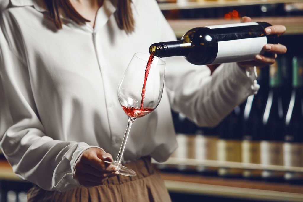 A sommelier's hands pouring red wine from a bottle into a glass in front of shelves of wine bottles