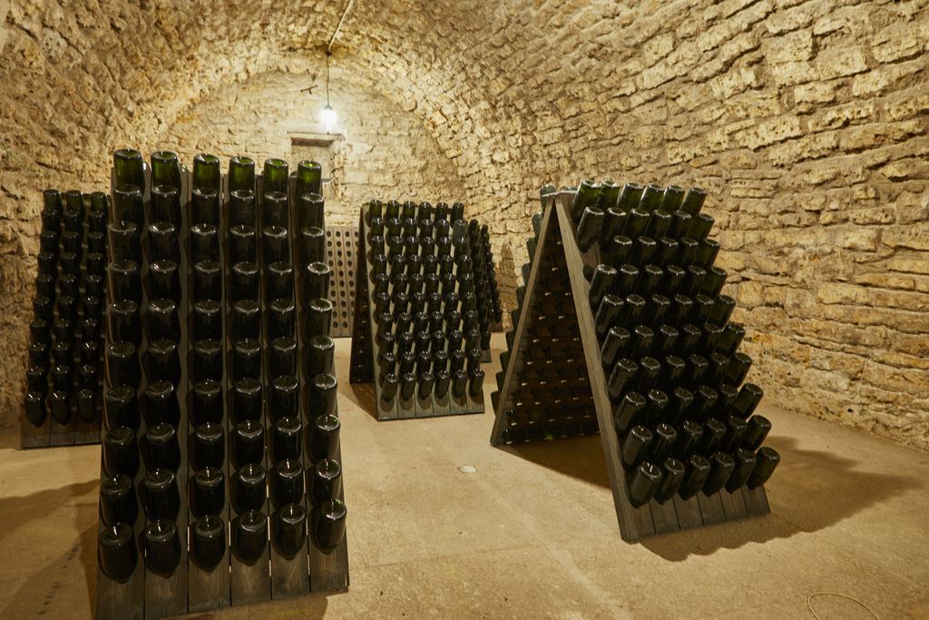Sparkling wine bottles stacked neck-down on wooden riddling racks inside a vaulted stone cellar