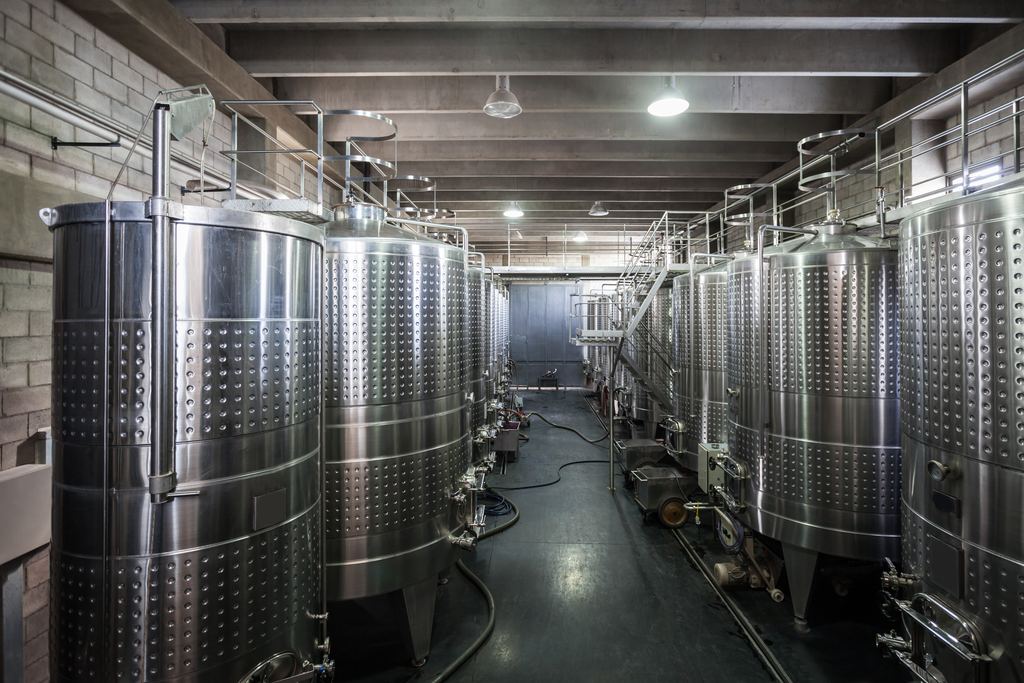Rows of stainless steel wine fermentation tanks with walkways and pipes inside a modern winery cellar