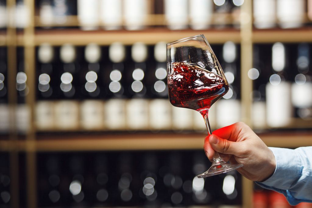 Hand swirling a glass of red wine against shelves of bottles in a wine cellar