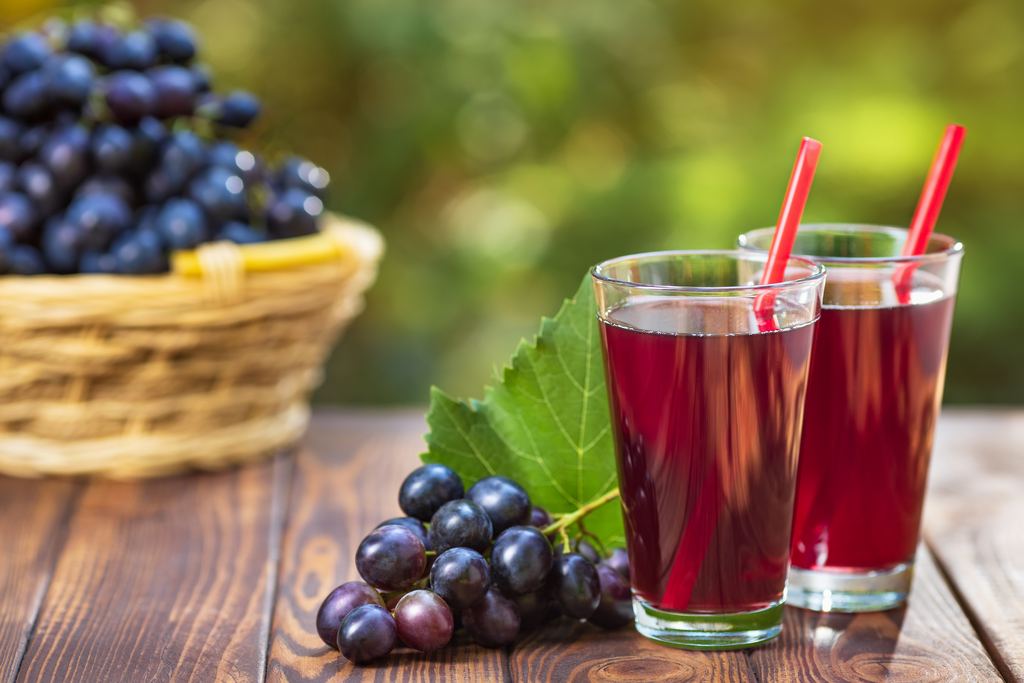 Two glasses of red grape juice with straws on a wooden table, next to black grapes and a wicker basket of grapes outdoors