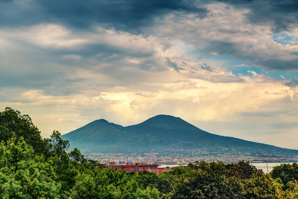 Mount Vesuvius under a dramatic sky, seen over the treetops near Naples