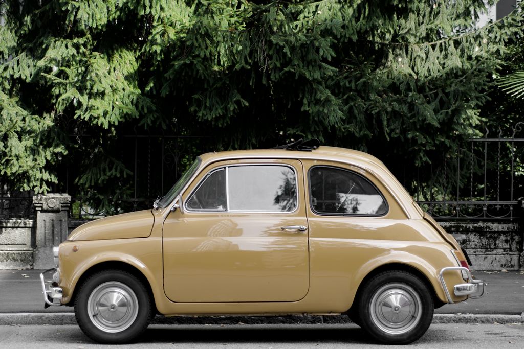 A beige vintage Fiat 500 parked under trees