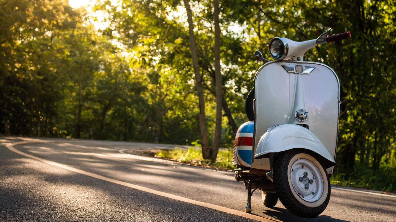 Classic white and blue vintage scooter parked on a quiet country road lined with trees in warm evening light
