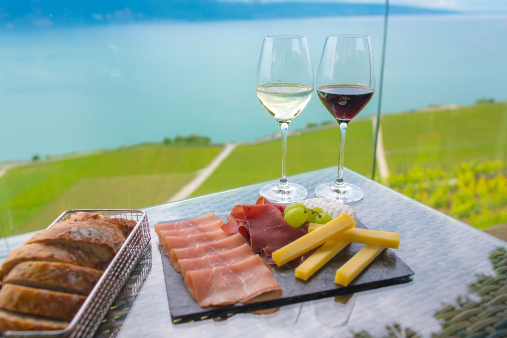 Glasses of white and red wine with cheese, cured ham and bread on a table overlooking vineyards and a lake